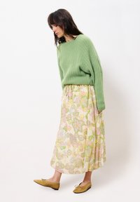 Grüner gerippter Pullover, floraler Maxirock in Pastellfarben, Ballerinas mit Schleifendetail. Weiche Texturen und fließende Silhouette stehen im Kontrast zum taillierten Oberteil.