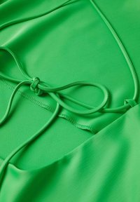 Haut en tissu vert avec des liens ajustables, une texture lisse et une finition mate, agrémenté de coutures soignées le long des bords.