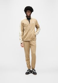 Μπεζ φόρμα από λείο ύφασμα, με μπουφάν με φερμουάρ και λευκές λεπτομέρειες, καθώς και ελαστικά μανίκια, συνδυασμένη με αντίστοιχο στενό παντελόνι και μαύρα αθλητικά παπούτσια.