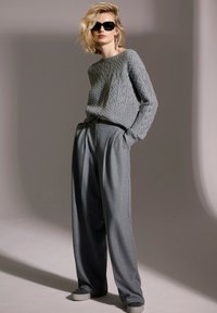 Grauer Kabelstrickpullover mit weitem Ausschnitt, kombiniert mit hochgeschnittenen grauen plissierten Hosen und grauen Slippern. Schwarze Sonnenbrille und Ohrringe.