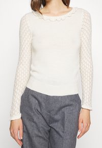 Dzianinowy top w jasnym kremowym kolorze z falbankowym dekoltem. Długie rękawy zdobione koronkowym wzorem. Miękka tekstura, dopasowany fason, zestawiony z szarymi spodniami.