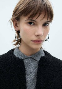 Giacca nera testurizzata, maglione grigio lavorato a maglia con scollo a V, orecchini a cerchio argentati e capelli corti castani in un ritratto ravvicinato.