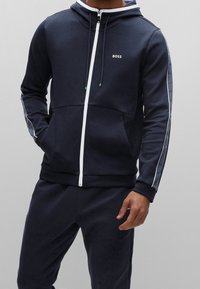 Marino hoodie gjord av mjukt tyg, har en fullständig framdraget dragkedja, snoddar och vita accentremsor med logotypdetaljer på ärmarna.