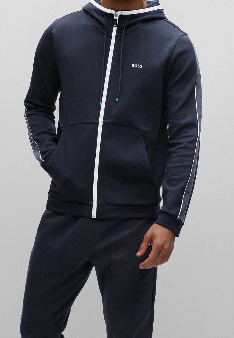 Marino hoodie gjord av mjukt tyg, har en fullständig framdraget dragkedja, snoddar och vita accentremsor med logotypdetaljer på ärmarna.