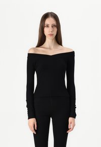 Svart off-shoulder topp med lange ermer, tettsittende design og subtil v-hals. Laget av myk, elastisk stoff. Kombinert med svarte bukser.
