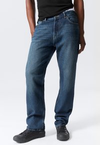 Jeans din denim albastru cu o croială relaxată, având un design clasic cu cinci buzunare, cusături galbene și închidere cu botton metalic.