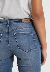 Femme portant un jean en denim bleu clair avec poches arrière et un haut blanc, montrant l’étiquette de la marque du jean sur la ceinture.