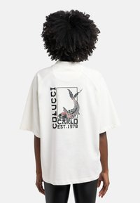 Witte oversized t-shirt met korte mouwen, voorzien van een grafische afbeelding van een koi-vis en de tekst "COLUCCI CALO EST. 1978" in zwart en rood. Gladde textuur.