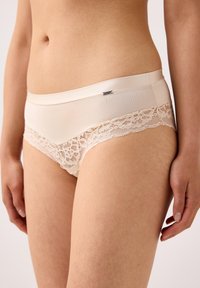 Beige alushamehkuilla varustetut alushousut, joissa on sileä kangas yläosassa ja herkkiä kukkalaceja helman kohdalla. Vyötäröllä on pieni metallinen logokoriste.