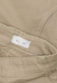 Beiger Baumwollstoff mit strukturiertem Finish, ausgestattet mit einem weißen Etikett, das mit einem Knopf gesichert ist, und detaillierten Nähten entlang der Kanten.