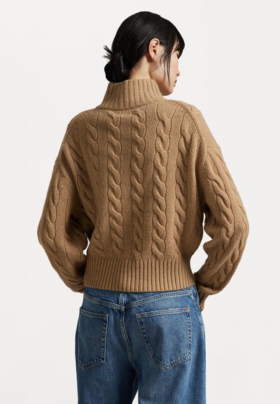 Maglione marrone a treccia con colletto alto, orlo a coste e motivo testurizzato. Abbinato a jeans blu, visto da dietro.