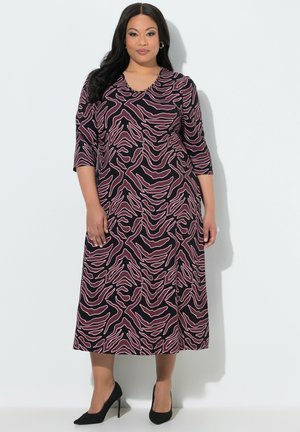 Femme portant une robe à rayures zèbre noires et roses avec des manches trois-quarts et des talons hauts noirs, debout devant un fond blanc.