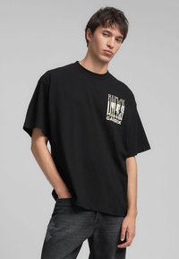 T-shirt nero a maniche corte in cotone con scollo rotondo. Presenta una stampa grafica bianca e gialla nella zona a sinistra del petto.
