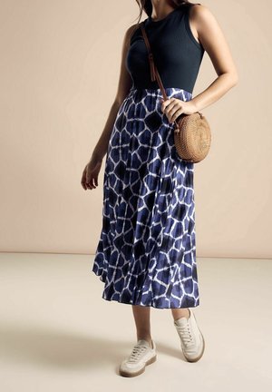 Kobieta ubrana w czarny top bez rękawów, midi spódnicę w niebiesko-biały wzór, białe trampki oraz okrągłą plecioną torebkę na ramię.