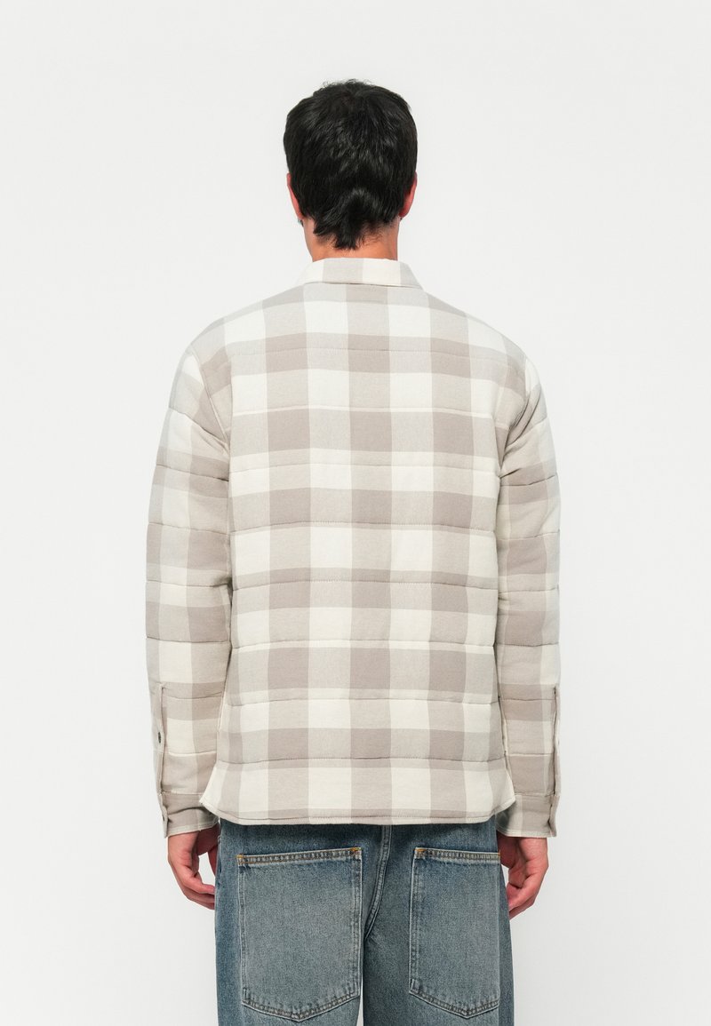 Mann in beiger und weiß karierter gepolsterter Jacke und verblassten blauen Jeans, der mit dem Rücken zu einer schlichten weißen Wand steht.