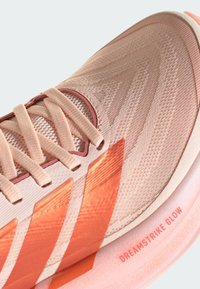 Gros plan sur une chaussure de course pêche et corail avec des lacets en tissu, une tige en mesh texturé et le texte "DREAMSTRIKE GLOW" sur la semelle intermédiaire.