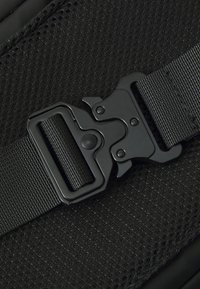 Černý nylonový popruh s plastovou sponou. Povrch má strukturovaný síťový design, který poskytuje odolnost a přilnavost.