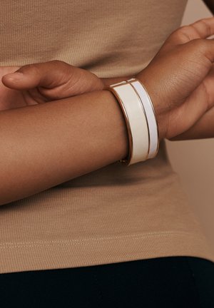 Gold-tone bangle bracelet features two white sections, one smooth and one with a textured line. Worn on a wrist against a beige top.