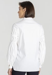 Kobieta z krótkimi brązowymi włosami, ubrana w białą koszulę z długimi rękawami, z marszczonymi detalami na rękawach, stojąca na jednolitym tle.