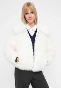 Giacca in pelliccia sintetica bianca con colletto alto, chiusura con zip e tasche laterali. Il modello indossa un maglione blu scuro sotto, posizionato su uno sfondo neutro.