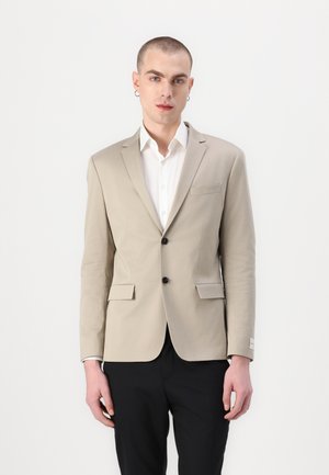 Blazer beige avec deux poches avant et deux boutons. Porté sur une chemise blanche, associé à un pantalon noir. Tissu lisse, revers crantés.