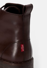 Tacón de bota de cuero marrón oscuro con tira de agarre, costuras visibles y una pequeña etiqueta de tela roja sobre un fondo blanco.