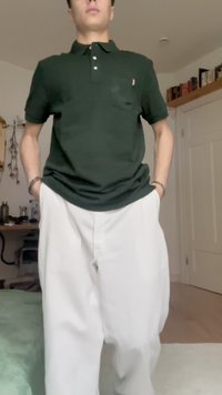 Grünes Baumwoll-Polohemd mit einer Brusttasche, kombiniert mit lockeren weißen Hosen. Der Hintergrund ist ein Innenraum mit hellen Wänden.