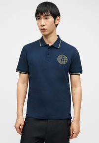 Νέος άνδρας που φοράει μπλε πόλο μπλούζα με κίτρινο περιθώριο και κυκλικό λογότυπο Versace Jeans Couture στο αριστερό στήθος, στέκεται μπροστά σε λευκό φόντο.