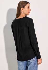Blusa nera a maniche lunghe con colletto rotondo, pieghe sottili sul retro e polsini con bottoni, abbinata a jeans in denim azzurro chiaro.