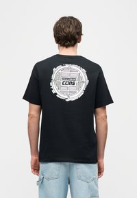 Άνδρας που στέκεται με την πλάτη γυρισμένη στην κάμερα, φοράει μαύρο t-shirt Converse CONS με λευκό κυκλικό γραφικό στο πάνω μέρος της πλάτης και ανοιχτόχρωμο μπλε τζιν.