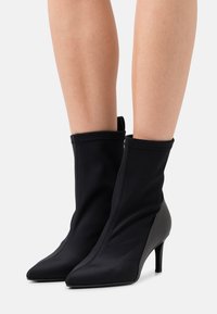 Ben som bär svarta, spetsiga ankelboots med smala klackar och ovandel i slät tyg mot en enkel vit bakgrund.