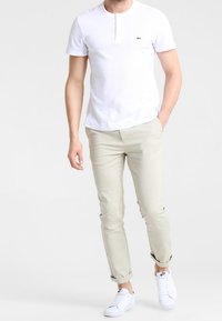 Maglietta henley bianca a maniche corte, pantaloni beige a gamba affusolata e sneakers bianche. Presenta un piccolo logo verde sulla maglietta.