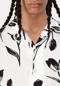 Camisa de manga corta en tejido blanco adornada con patrones florales negros, con cuello en pico y un cierre de botón único.