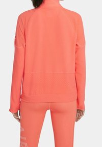 Chaqueta deportiva coral con un cuello alto, tejido texturizado y detalles perforados en las mangas, combinada con leggings ajustados a juego.