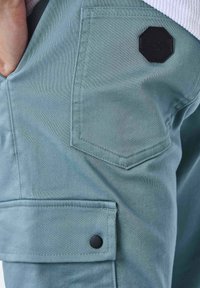 Pantaloni cargo blu chiaro con finitura opaca, dotati di tasca laterale e di una toppa ottagonale nera. Tessuto liscio, design funzionale.