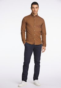 Camicia marrone con bottoni e colletto, abbinata a pantaloni blu scuro e scarpe da ginnastica bianche. Tessuto liscio, design aderente e trama sottile.