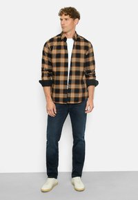 Kariertes Hemd in Schwarz, Orange und Blau, mit Button-Down-Kragen, hochgekrempelten Ärmeln, getragen über einem weißen T-Shirt und kombiniert mit dunklen Jeans und weißen Schuhen.