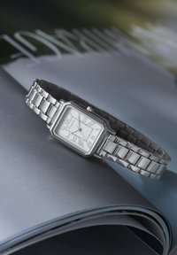 Montre rectangulaire en argent avec chiffres romains sur un cadran blanc et bracelet métallique à maillons, posée sur un magazine ouvert brillant.