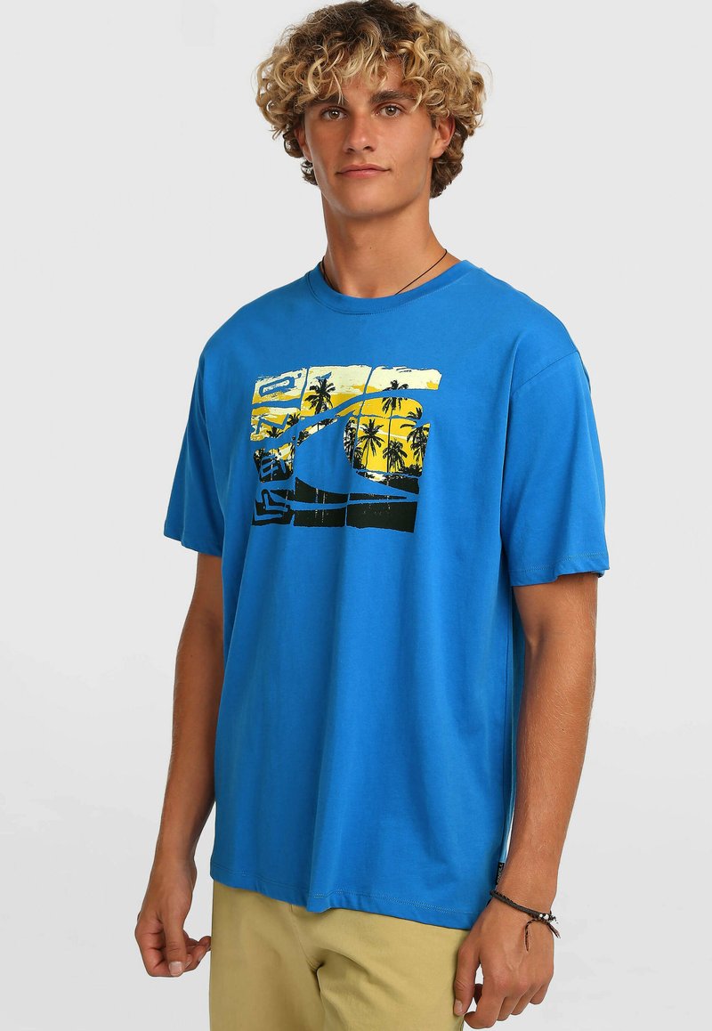 T-shirt en coton bleu arborant une impression graphique avec des accents jaunes et noirs, représentant des palmiers et un motif de vagues. Manches courtes, col rond.
