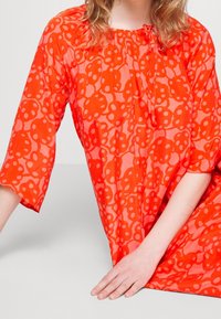 Robe rouge à motifs avec manches bouffantes, dotée d'un col froncé et de motifs floraux. Tissu léger et semi-transparent.