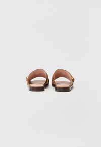 Brune lær sandaler med åpen bak, flate såler og en dekorativ spenne på siden. Den indre foringen er i en lys beige farge.