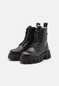 Schwarze Leder-Stiefeletten mit einer dicken schwarzen Sohle, sechseckigem Muster und einem Kunstfellfutter am oberen Rand. Merkmale sind die Schnürung vorne und eine Zuglasche.