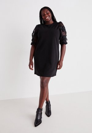 Schwarzes Kleid mit kurzen, puffartigen Spitzenärmeln. Glatter Stoff mit lässigem Schnitt und rundem Halsausschnitt. Ein Paar schwarze Stiefeletten.