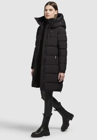 Abrigo de plumas negro con capucha, cierre frontal y un diseño acolchado prominente. Lleva encima un suéter negro y leggings, combinado con botas negras.