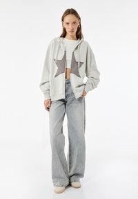 Lichtgrijze zip-up hoodie met twee donkergrijze ster applicaties, gecombineerd met hoge, losvallende lichtblauwe jeans. Gelaagd stof.