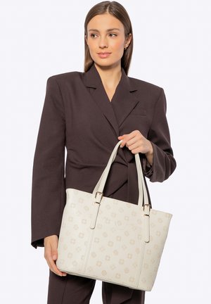 Cremefarbene Tragetasche mit strukturierter Oberfläche, goldfarbenen Beschlägen und Blumenmustern, gehalten von einer Person in einem dunklen Blazer.