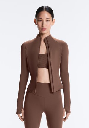 Ženska z temnimi lasmi nosi rjavo jakno z zadrgo preko ujemajočega se športnega modrčka in visokih legic, stoji pred enobarvnim svetlim ozadjem.