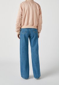 Personne portant une veste bomber rose clair et un jean bleu taille haute à jambes larges, debout de dos sur un fond uni.