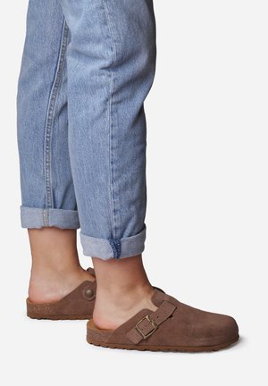 Zuecos de ante marrón con correa ajustable y hebilla de metal, combinados con jeans de mezclilla azul claro enrollados que presentan un ajuste relajado.