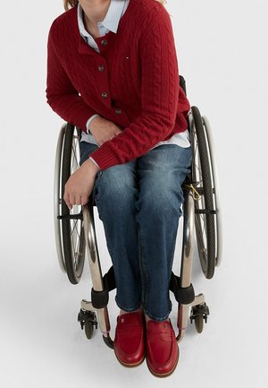 Persona con jeans blu, maglione rosso e scarpe rosse seduta su una sedia a rotelle manuale con le mani appoggiate sulla ruota, su uno sfondo neutro.